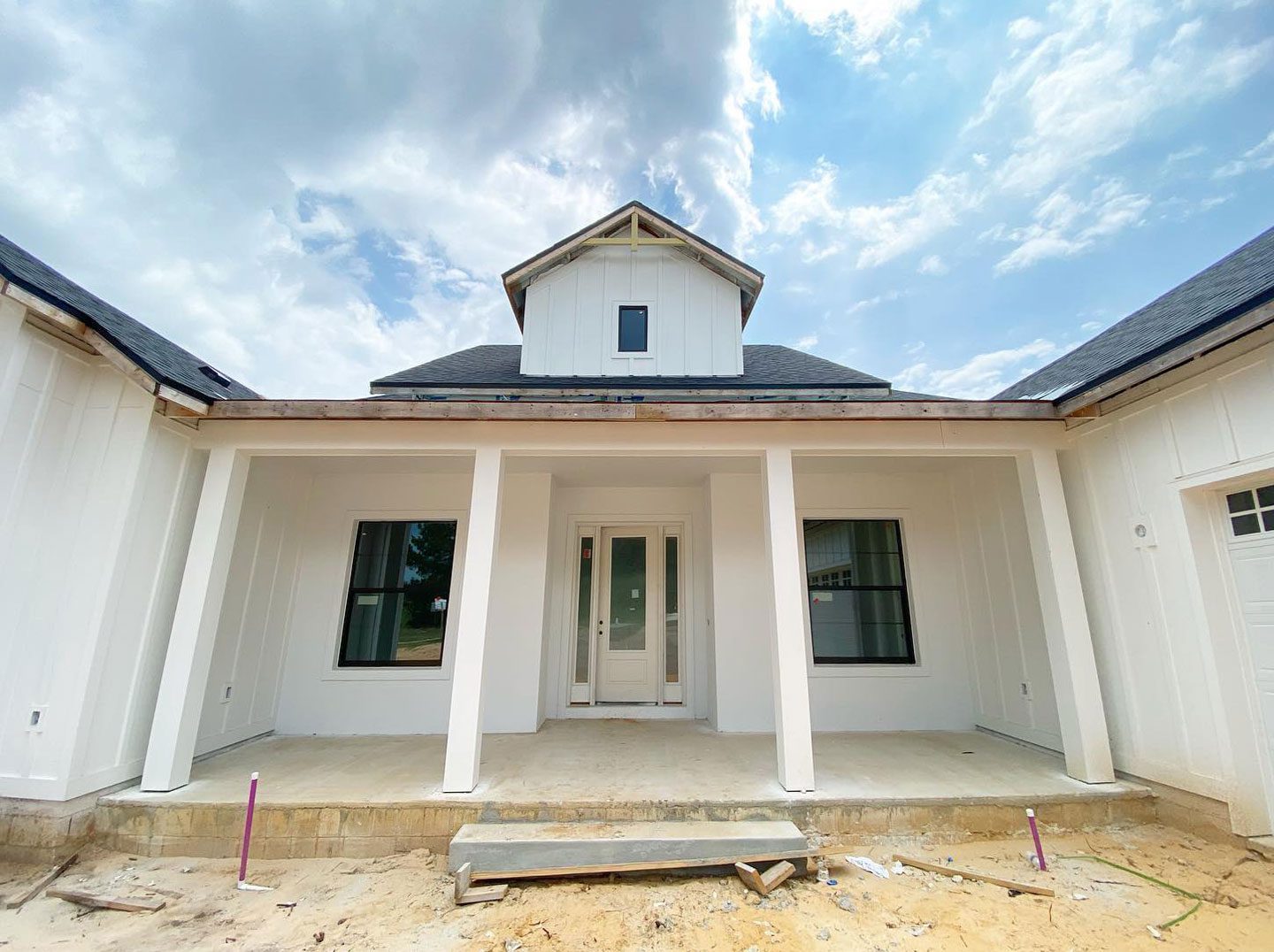 A white house under construction with a covered front porch and two windows on either side of the door. The house has a gabled roof and bright sky with clouds overhead.