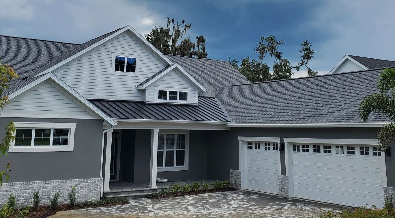 A modern grey home with a gable roof, white trim, and a two-car garage. The front yard has a brick driveway and minimal landscaping. A partially cloudy sky is in the background.