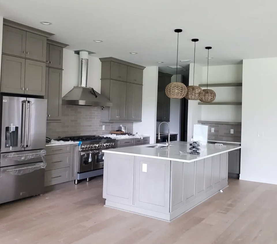 Modern kitchen with gray cabinets, stainless steel appliances, a large island with white countertop, and three pendant lights. Wooden floor and light gray subway tile backsplash.