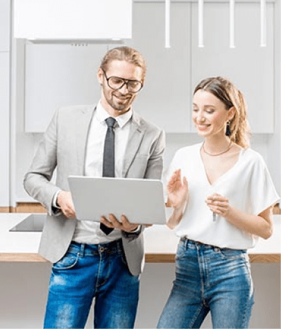 Two people standing in a modern kitchen, looking at a laptop. The man is wearing a suit and the woman is in casual attire. Both appear engaged in conversation.