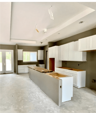 A partially constructed kitchen with white cabinets, a central island, and light gray walls. The floor is unfinished, and there are exposed wires hanging from the ceiling.