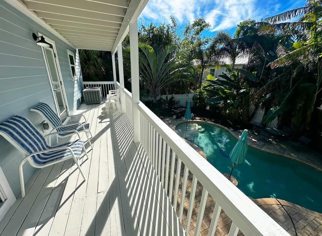 A white porch overlooks a backyard pool surrounded by palm trees. Two striped lounge chairs are on the porch.